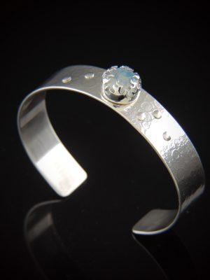 A half-inch wide sterling silver cuff bracelet featuring a textured, roll-printed surface and small silver rivet accents. Centered on the band is a round, iridescent rainbow moonstone secured by an intricate, crown-like flame bezel, displayed against a reflective black background.