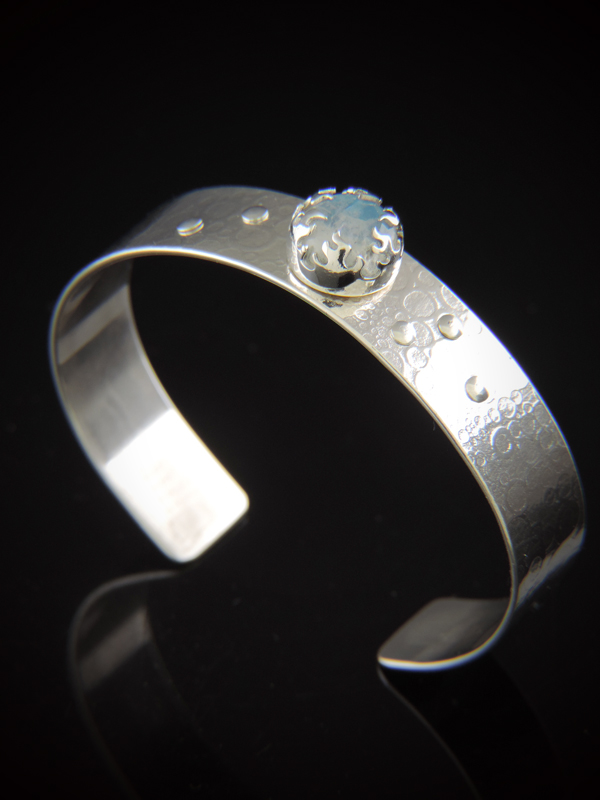 A half-inch wide sterling silver cuff bracelet featuring a textured, roll-printed surface and small silver rivet accents. Centered on the band is a round, iridescent rainbow moonstone secured by an intricate, crown-like flame bezel, displayed against a reflective black background.
