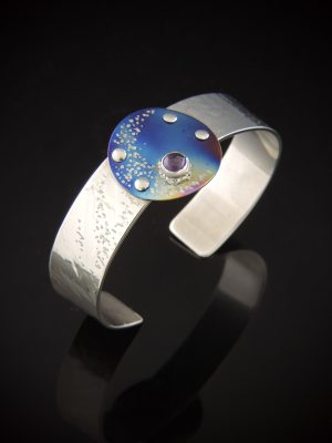 Sugar and Spice artisan cuff bracelet featuring a half-inch roll-printed sterling silver band with a starry texture, topped with a circular torch-fired blue titanium disk, silver rivets, and a purple amethyst cabochon.
