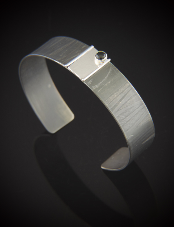 Black Martini Niobium with Sterling Silver and Black Tourmaline Cuff. Half inch wide niobium cuff features a torch fired charcoal color finish with a roll printed texture. Accented with a half inch by one inch sterling silver background framed with sterling silver and a 4mm black tourmaline stone set in fine silver. Approximate size: S (fits wrist sizes 6.5 inches)