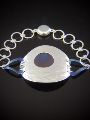 A contemporary sterling silver bracelet featuring a large, textured shield-shaped centerpiece with a circular blue titanium inlay, connected by blue triangular accents to a silver link chain with a magnetic clasp.A contemporary sterling silver bracelet featuring a large, textured shield-shaped centerpiece with a circular blue titanium inlay, connected by blue triangular accents to a silver link chain with a magnetic clasp.