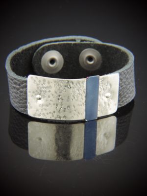 A wide dark gray pebbled leather cuff bracelet featuring a rectangular sterling silver plate riveted to the center. The silver has a hammered, dimpled texture and is accented by a thin, vertical stripe of smooth blue titanium in the middle. The cuff is shown on a reflective black surface with two silver snaps visible in the interior.
