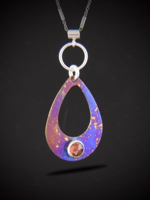 Handcrafted open teardrop pendant made of torch-fired titanium in vibrant blue and purple hues, featuring a red rose-cut garnet bezel-set at the bottom, suspended from a black rhodium satellite chain.
