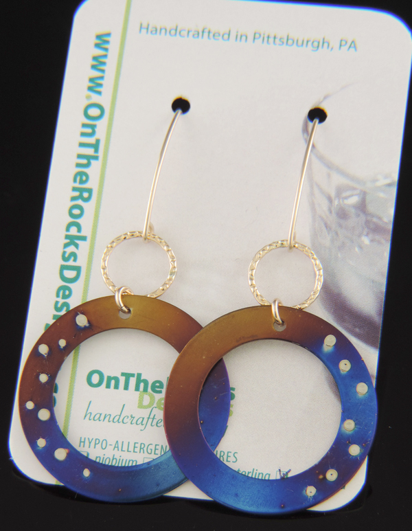 A pair of circular titanium dangle earrings on a white card. The disks feature a torch-fired gradient of blue, purple, and gold with small silver weld spots resembling stars. They are attached via gold links to long, gold-filled V-shaped ear wires.