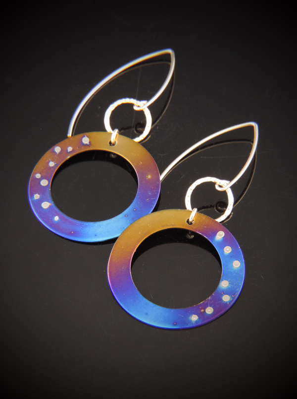 A pair of circular titanium dangle earrings on a black reflective surface. The disks feature a torch-fired gradient of blue, purple, and gold with small silver weld spots resembling stars. They are attached via gold links to long, gold-filled V-shaped ear wires.