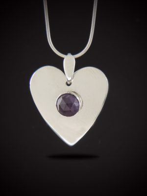 A heart-shaped sterling silver pendant featuring a central round rose-cut purple amethyst, suspended from a silver snake chain against a black background.