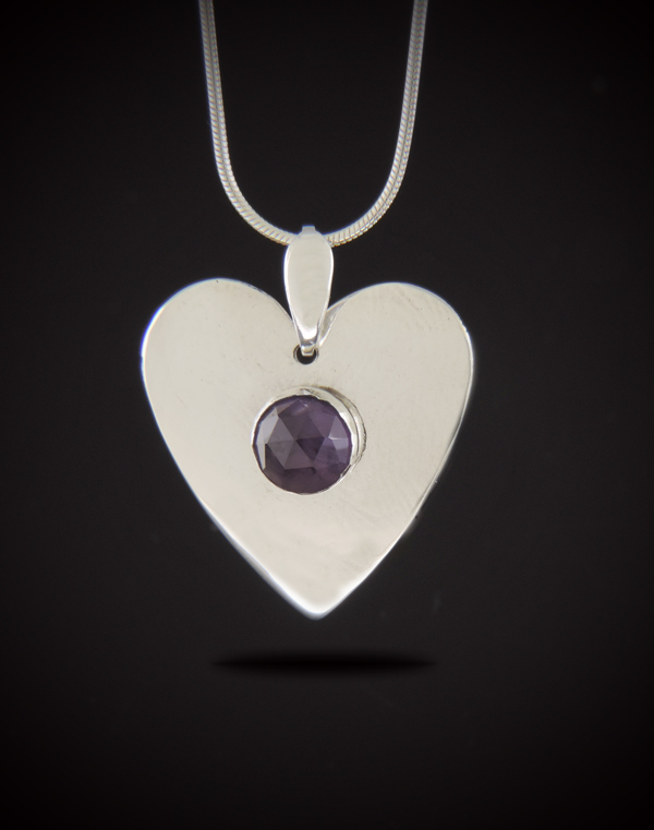 A heart-shaped sterling silver pendant featuring a central round rose-cut purple amethyst, suspended from a silver snake chain against a black background.