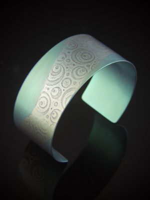A wide, one-inch titanium cuff bracelet featuring a hand-anodized apple green and light lime finish. The metal is roll-printed with a pattern of varying concentric circles and dots. The cuff is shown reflecting on a dark, polished surface, highlighting its vibrant iridescent color and curved silhouette.