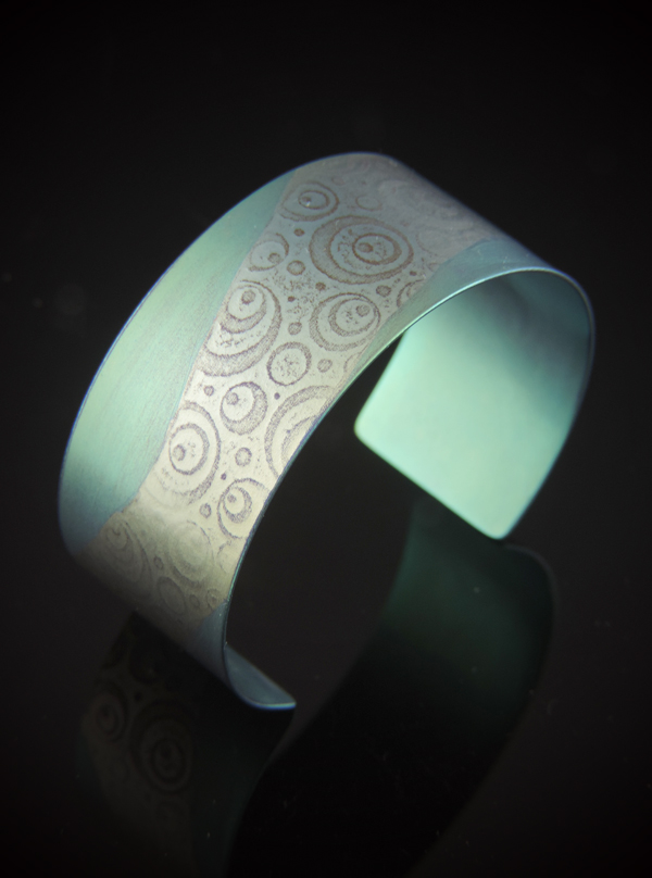 A wide, one-inch titanium cuff bracelet featuring a hand-anodized apple green and light lime finish. The metal is roll-printed with a pattern of varying concentric circles and dots. The cuff is shown reflecting on a dark, polished surface, highlighting its vibrant iridescent color and curved silhouette.
