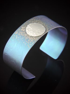 A one-inch wide teal-anodized titanium cuff bracelet featuring a circular sterling silver disc accent off-center. The teal surface is embellished with a scattered, shimmering texture that resembles stardust. The cuff is photographed on a black reflective surface, showcasing its vibrant color and the contrast between the silver and teal metals.