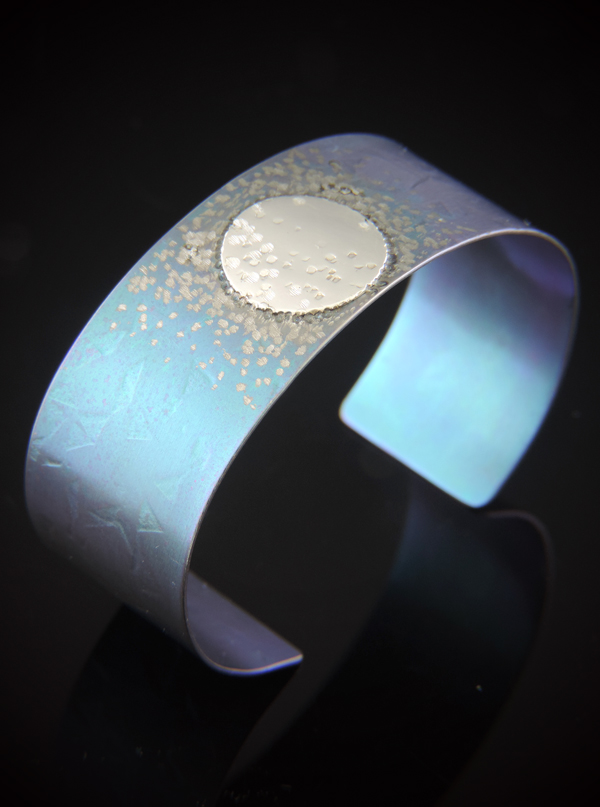 A one-inch wide teal-anodized titanium cuff bracelet featuring a circular sterling silver disc accent off-center. The teal surface is embellished with a scattered, shimmering texture that resembles stardust. The cuff is photographed on a black reflective surface, showcasing its vibrant color and the contrast between the silver and teal metals.