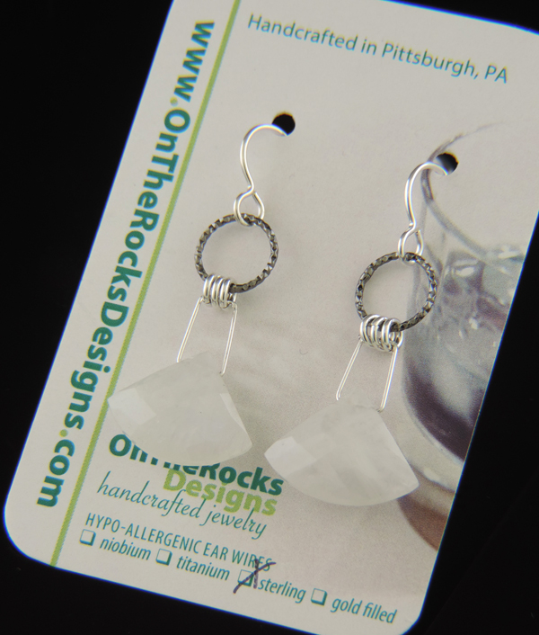 A pair of triangle-shaped, milky white faceted moonstone earrings hanging from textured sterling silver rings and silver earwires on a reflective black surface.