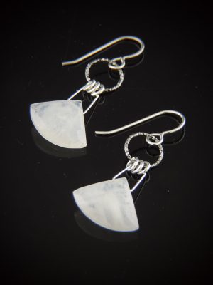 A pair of triangle-shaped, milky white faceted moonstone earrings hanging from textured sterling silver rings and silver earwires on a reflective black surface.