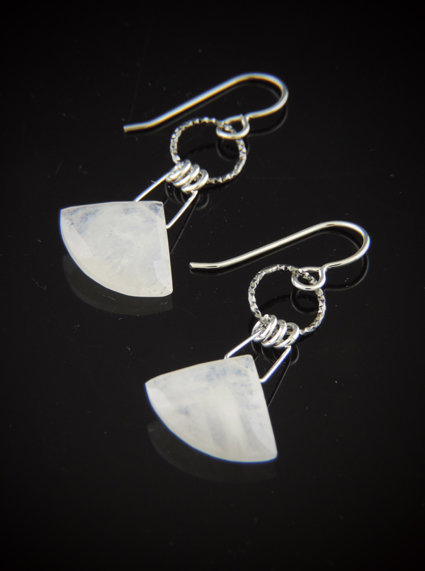 A pair of triangle-shaped, milky white faceted moonstone earrings hanging from textured sterling silver rings and silver earwires on a reflective black surface.