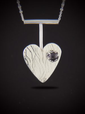 A handcrafted sterling silver heart-shaped pendant with a rippled, roll-printed texture. The heart features a small, round purple amethyst cabochon set in a decorative silver flame bezel. The pendant hangs from a modern T-bar style drop on a silver chain against a dark background.