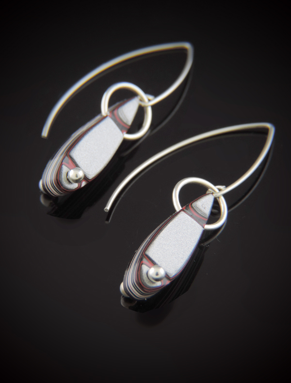Pair of teardrop-shaped Fordite earrings with shimmery silver and red paint layers. The stones are suspended by sterling silver rings and feature a small silver bead at the bottom, hanging from long V-shaped sterling silver ear wires against a black reflective background.