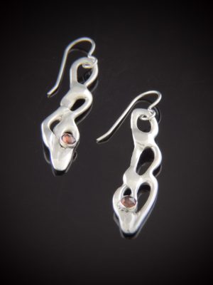 Handcrafted sterling silver dangle earrings with a fluid, organic cutout design and a small red rose-cut garnet gemstone at the base, shown on a reflective black surface.