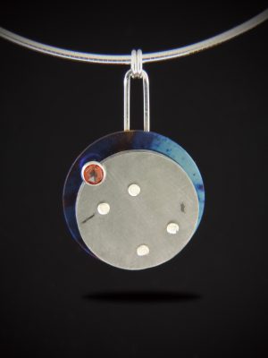 A circular mixed-metal pendant featuring a textured Gibeon meteorite disk layered over a blue-toned torch-fired titanium base. A small red rosecut garnet is set near the top, and the piece is held by a tall sterling silver bail on an omega chain against a black background.