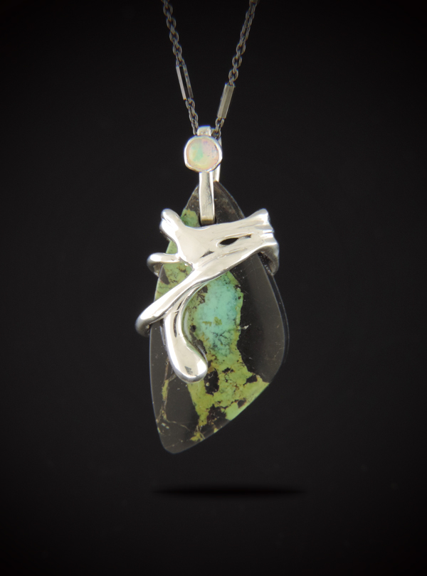 A teardrop-shaped Black Jack Turquoise pendant with a dark matrix and green-blue veins, wrapped in fluid sterling silver with a small opal set in the bail, hanging from a dark rhodium chain against a black background.