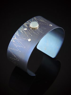 Handcrafted blue titanium cuff bracelet with an etched mountain design, featuring a central faceted blue-green aquamarine gemstone and fine silver rivet details against a black reflecting background.