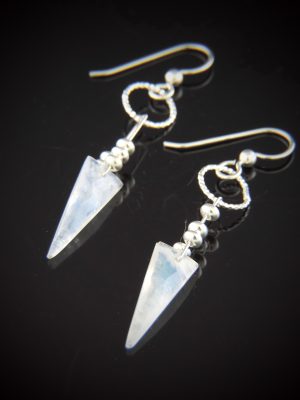 A pair of Moonjuice Moonstone Dangle Earrings on a reflective black surface, featuring sharp, spear-shaped rainbow moonstones, sterling silver saucer beads, and a textured 10mm round link hanging from 20g silver earwires.