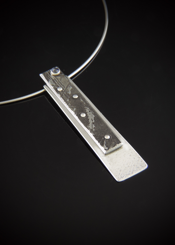 Angled close-up of the Blue Moon meteorite and sterling silver pendant resting on a reflective black surface, highlighting its layered dimension, metallic patterns, and rose-cut blue sapphire.