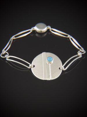 A sterling silver bracelet on a reflective black surface, featuring a textured 1-inch central disk accented by a small blue turquoise stone, connected to a paperclip link chain with a smooth round magnetic clasp.