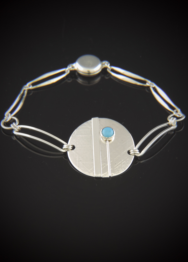 A sterling silver bracelet on a reflective black surface, featuring a textured 1-inch central disk accented by a small blue turquoise stone, connected to a paperclip link chain with a smooth round magnetic clasp.