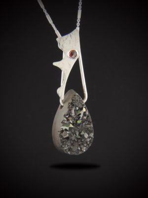 A vertical statement necklace named Widow's Kiss, featuring a heavy teardrop-shaped black druzy gemstone with a highly textured, sparkling crystalline front surface. Above the druzy hangs an organic, hand-sculpted textured sterling silver accent piece holding a small, round, crimson-red rose-cut garnet gemstone. The pendant is suspended from a delicate sterling silver chain against a solid black background.