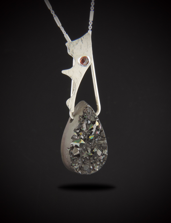 A vertical statement necklace named Widow's Kiss, featuring a heavy teardrop-shaped black druzy gemstone with a highly textured, sparkling crystalline front surface. Above the druzy hangs an organic, hand-sculpted textured sterling silver accent piece holding a small, round, crimson-red rose-cut garnet gemstone. The pendant is suspended from a delicate sterling silver chain against a solid black background.