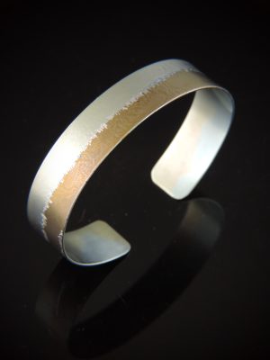 A half-inch wide titanium cuff bracelet displayed on a reflective black background. The bracelet features a two-tone anodized finish, split lengthwise with a soft metallic kiwi green color on one side and a warm gold tone on the other. A jagged, organic hand etched textured line runs directly down the center where the two colors meet.