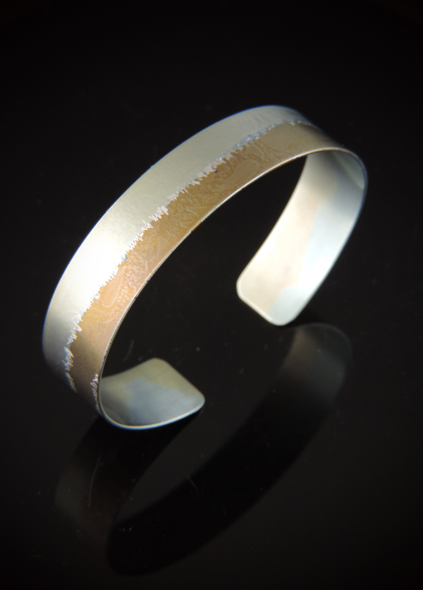 A half-inch wide titanium cuff bracelet displayed on a reflective black background. The bracelet features a two-tone anodized finish, split lengthwise with a soft metallic kiwi green color on one side and a warm gold tone on the other. A jagged, organic hand etched textured line runs directly down the center where the two colors meet.