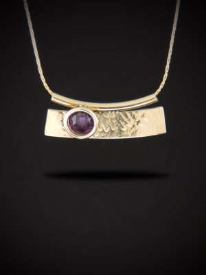 Handcrafted gold-filled bar pendant necklace featuring an organic roll-printed texture and a bezel-set 6mm rose-cut purple amethyst. The curved pendant hangs seamlessly from a sleek gold-filled Cardano chain passing through a matching tube bail, displayed against a dark gradient background.