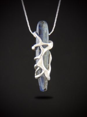 Brooklynite pendant necklace featuring a slender blue kyanite stone wrapped in an organic sterling silver cast setting, suspended from a sterling silver snake chain against a dark background.
