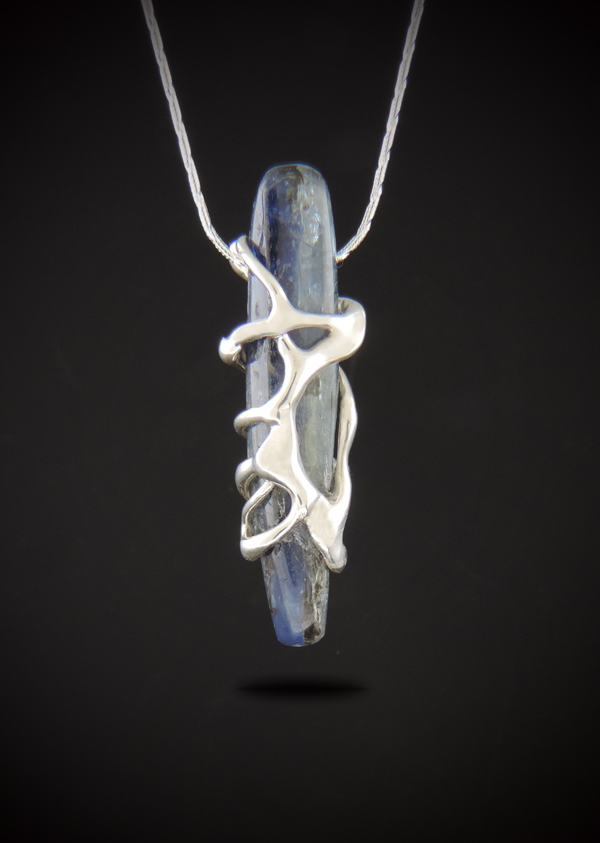 Brooklynite pendant necklace featuring a slender blue kyanite stone wrapped in an organic sterling silver cast setting, suspended from a sterling silver snake chain against a dark background.